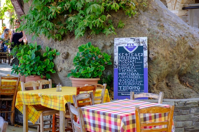 des tables colorées d'une taverne grecque, sur une rue piétonne, avec un écriteau où sont inscrits des plats écrits en grec